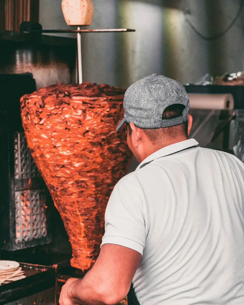 A cook slicing meat from a trompo of pork al pastor
